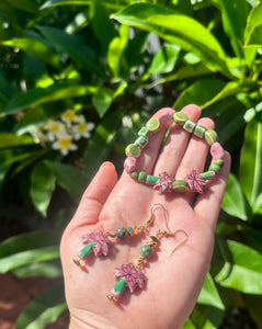 Pink &amp; green ceramic palm tree earrings with matching bracelet on hand with frangipani tree behind