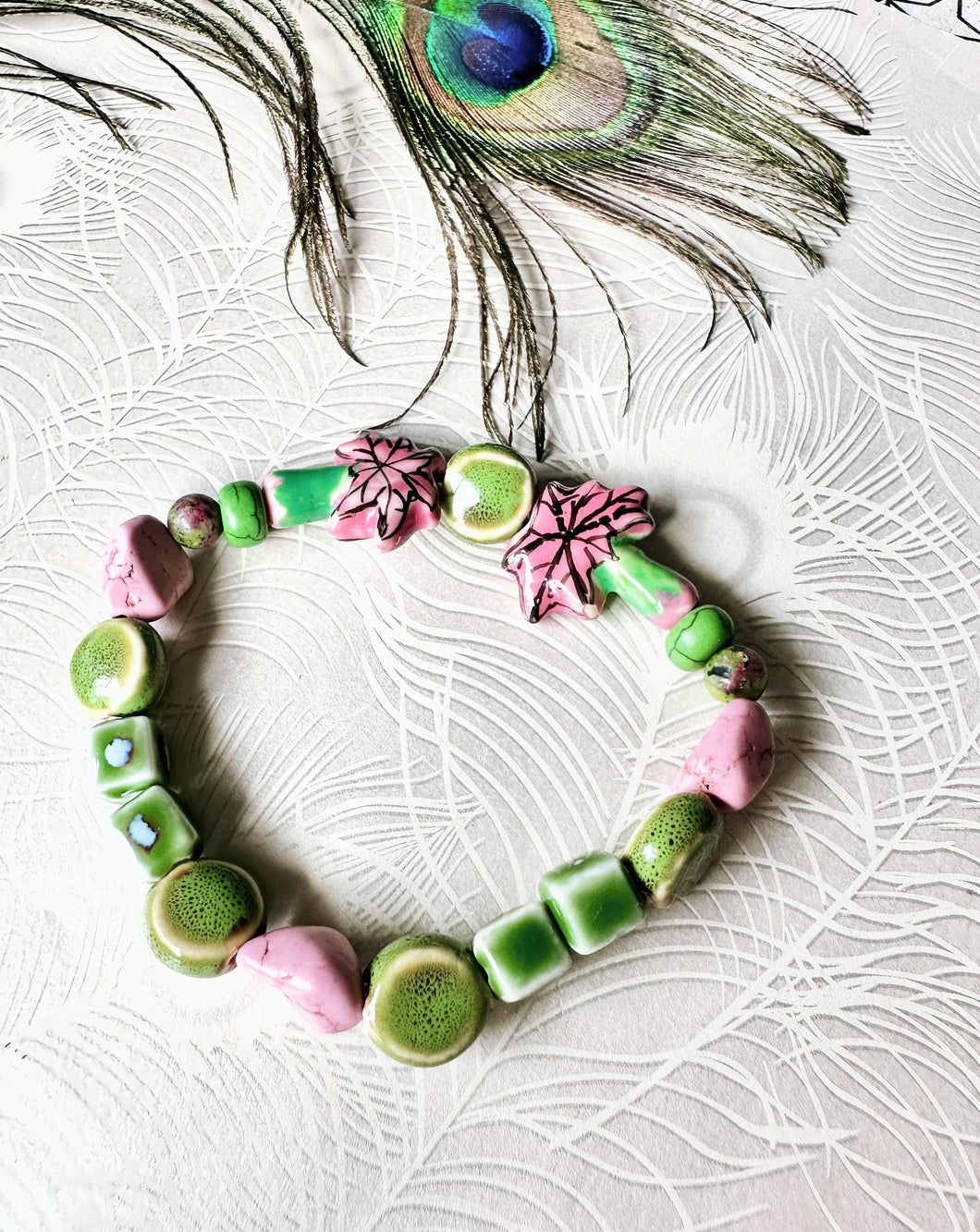 Pink & green ceramic palm tree beaded bracelet on peacock patterend backgound with peacock feather