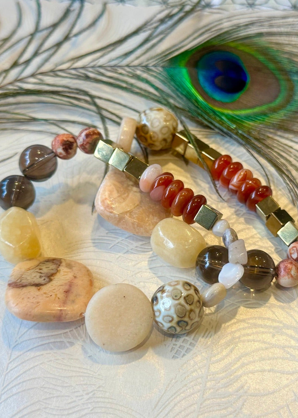  two Carnelian, peach stone, Smokey quartz, hematite gemstone & ceramic beaded bracelet on a peacock feather paper background with a peacock feather above