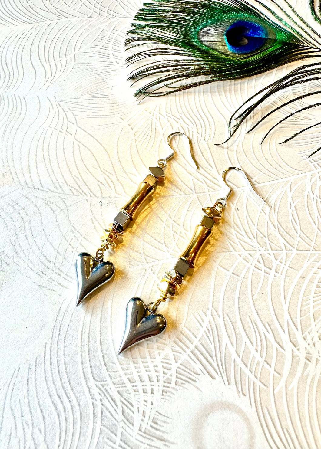Silver heart earrings with gold and hematite beads next to a peacock feather 