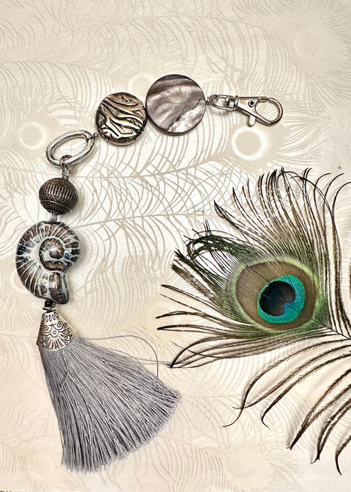 Keychain bag tag with silver gray tassel beads and ceramic shell feature bead sitting near a peacock feather 