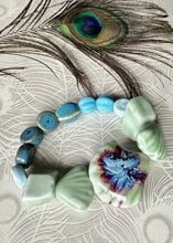 Load image into Gallery viewer, Bracelet with Ceramic shell shaped large center bead, and multi shaped surrounding beadss on a peacock background with peacock featherpale green in colour