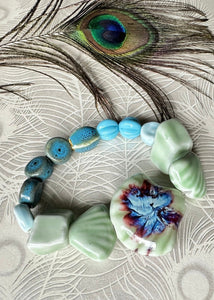 Bracelet with Ceramic shell shaped large center bead, and multi shaped surrounding beadss on a peacock background with peacock featherpale green in colour