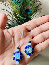 Load image into Gallery viewer, Iridescent Cobalt blue round enamel cloissone flower design earrings including Swarovski crystal &amp; sterling silver textured long ear hooks sitting on a hand with a white background with peacock feathers