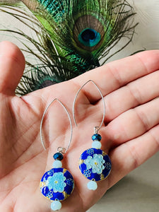 Iridescent Cobalt blue round enamel cloissone flower design earrings including Swarovski crystal & sterling silver textured long ear hooks sitting on a hand with a white background with peacock feathers