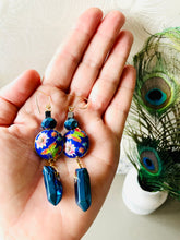 Load image into Gallery viewer, Iridescent Cobalt blue raw quartz pointed stone earrings with round enamel flower &amp; butterfly design, including Swarovski crystal &amp; 14ct gold filled ear hooks sitting on hand over a white background with a small vase filled with peacock feather