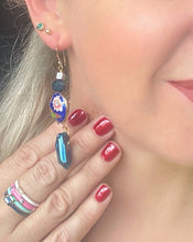 Load image into Gallery viewer, Iridescent Cobalt blue raw quartz pointed stone earrings with round enamel flower &amp; butterfly design, including Swarovski crystal &amp; 14ct gold filled ear hooks worn on a model with blond hair holding up her hand with red nails &amp; coloured enamel stacking rings