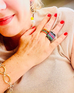 a range of 12 different coloured enamel stacking rings worn on  fingers with red nail polish and yellow earrings with a gold bracelet and beige t-shirt