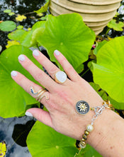 Load image into Gallery viewer, gold engraved coiled snake ring with blue and clear CZ stones worn on index finger with gold mother of pearl ring worn on ring finger &amp; bracelet with bronze pearls, black starburst enamel charm on wrist set against a lilly pond