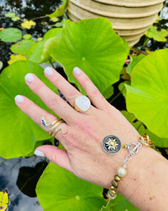 gold engraved coiled snake ring with blue and clear CZ stones worn on index finger with gold mother of pearl ring worn on ring finger & bracelet with bronze pearls, black starburst enamel charm on wrist set against a lilly pond
