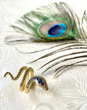 Load image into Gallery viewer, gold engraved coiled snake ring with blue and clear CZ stones against a white background with peacock feather
