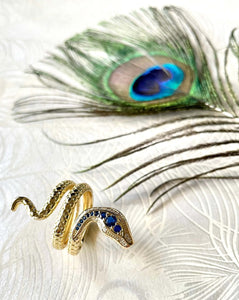 gold engraved coiled snake ring with blue and clear CZ stones against a white background with peacock feather