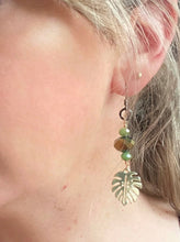 Load image into Gallery viewer, Gold-metal-monstera-leaf-with-olive-green-Czech-crystal-beads-gunmetal-ear-hooks-on- blonde-haired-model