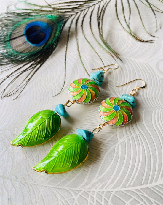 Iridescent Green leaf & round enamel cloissone flower design earrings including turquoise stones & 14ct gold filed ear hooks sitting on white paper with peacock feather