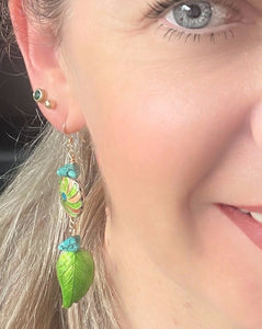 Iridescent Green leaf & round enamel cloissone flower design earrings including turquoise stones & 14ct gold filed ear hooks worn on a model with blonde hair & blue eyes