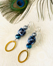 Load image into Gallery viewer, Cobalt blue cube ceramic bead earrings with Swarovski &amp; Czech crystal, a gold toned oval feature &amp; sterling silver ear hooks on peacock patterned paper background with feather