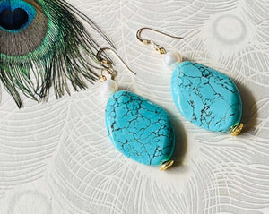 Large turquoise oval gemstone earring with small freshwater white pearl and gold ear hooks against a white background with a peacock feather