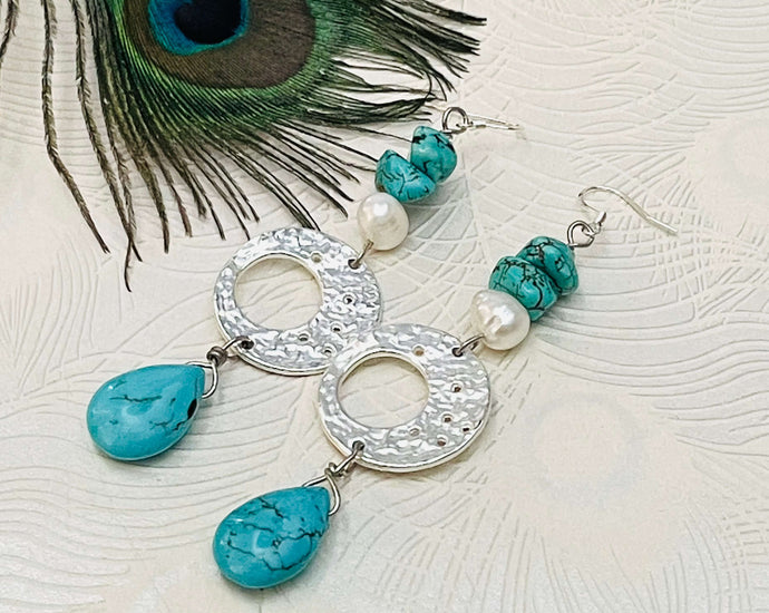 Earrings with a Turquoise teardrop shape stone hanging from silver disc with pearl & turquoise stone on a silver ear hook against a white background with a peacock feather