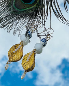 White ceramic beads with blue flowers, white Czech faceted crystals and gold metal leaf on 14ct filled gold ear hooks sitting on mirror with sky background & peacock feather