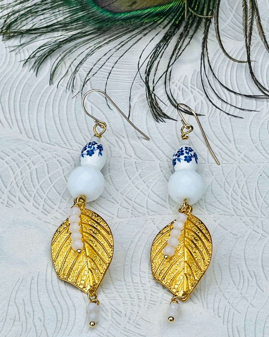 White ceramic beads with blue flowers, white Czech faceted crystals and gold metal leaf on 14ct filled gold ear hooks sitting on paper patterned background with peacock feather