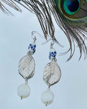 Load image into Gallery viewer, White ceramic beads with blue flowers, crystals and mothers of pearl beads with silver  metal leaf on sterling silver ear hooks sitting on white background with peacock feather