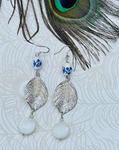 Load image into Gallery viewer, White ceramic beads with blue flowers, crystals and mothers of pearl beads with silver  metal leaf on sterling silver ear hooks sitting on patterned paper background with peacock feather