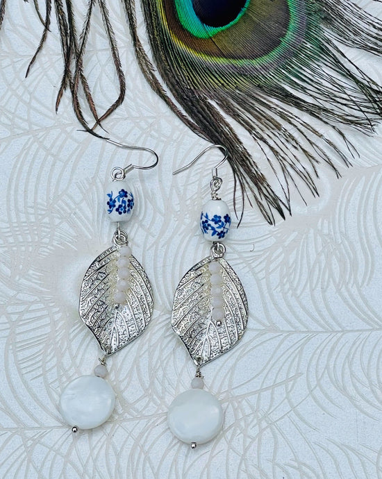 White ceramic beads with blue flowers, crystals and mothers of pearl beads with silver  metal leaf on sterling silver ear hooks sitting on patterned paper background with peacock feather