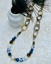 Load image into Gallery viewer, Necklace with blue &amp; white ceramic flower beads, gold hematite  &amp; cobalt blue &amp; white Czech crystal beads with a silver clasp with gold &amp; silver toned chain sitting on a patterned paper background with peacock feathers in a vase