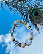 Load image into Gallery viewer, Bracelet with genuine coin shaped Keshi pearl feature bead, blue &amp; white ceramic flower beads, freshwater pearls &amp; gold hematite beads with a silver clasp with gold &amp; silver toned chain on paper patterned backgound with peacock feather  n mirrored surface with sky &amp; peacock feather