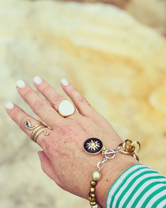 Gold restatement ring with mother of pearl inlay & gold snake ring both worn on a hand against a sand background with a bracelet of pearl & black enamel charm