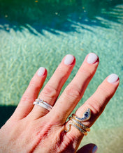 Load image into Gallery viewer, gold engraved coiled snake ring with blue and clear CZ stones worn on index finger with white nails against a turquiose pool background with white enamel stacking rings on ring finger