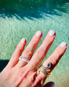 gold engraved coiled snake ring with blue and clear CZ stones worn on index finger with white nails against a turquiose pool background with white enamel stacking rings on ring finger