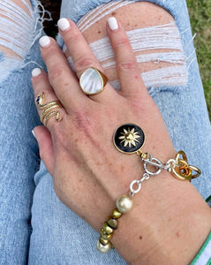 Gold restatement ring with mother of pearl inlay & gold snake ring both worn on a hand against a pair of ripped jeans with a bracelet of pearl & black enamel charm