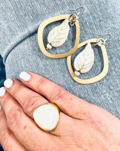 Gold restatement ring with mother of pearl inlay  worn on a hand against a pair of blue jeans with a pair of gold & white enamel leaf earrings