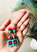 Load image into Gallery viewer, Turquoise square &amp; red crystal earrings with red enamel cloisonné bead &amp; 14ct gold filled ear hooks on a hand with beige background with peacock feathers in a small vase