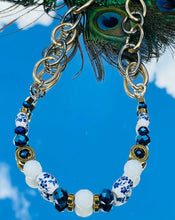 Load image into Gallery viewer, Necklace with blue &amp; white ceramic flower beads, gold hematite  &amp; cobalt blue &amp; white Czech crystal beads with a silver clasp with gold &amp; silver toned chain on a sky background with peacock feather