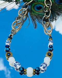 Necklace with blue & white ceramic flower beads, gold hematite  & cobalt blue & white Czech crystal beads with a silver clasp with gold & silver toned chain on a sky background with peacock feather