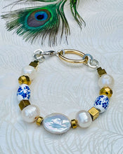 Load image into Gallery viewer, Bracelet with genuine coin shaped Keshi pearl feature bead, blue &amp; white ceramic flower beads, freshwater pearls &amp; gold hematite beads with a silver clasp with gold  &amp; silver toned chain on paper patterned backgound with peacock feather