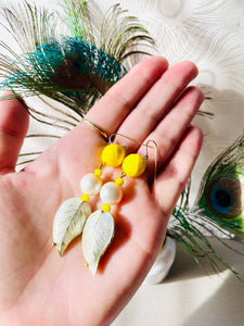 Iridescent white enamel leaf cloissone earrings with freshwater pearl, yellow faceted crystal & 14ct gold filled ear hooks sitting on a hand in front of a bunch of peacock feathers